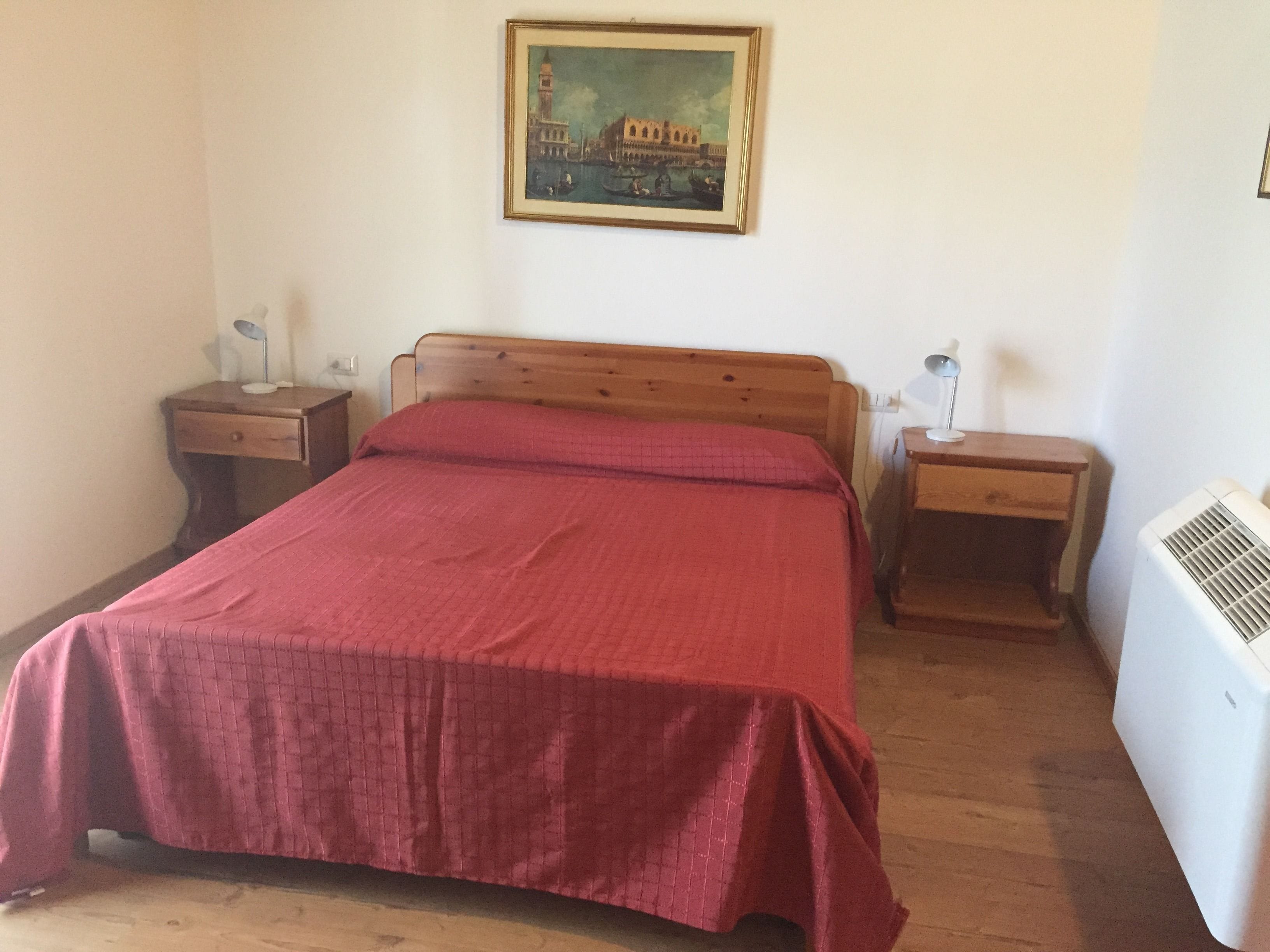 A bedroom with a wooden bed, a red bedspread, two bedside tables with lamps, a wall-mounted air conditioner, and a framed painting of a cityscape.