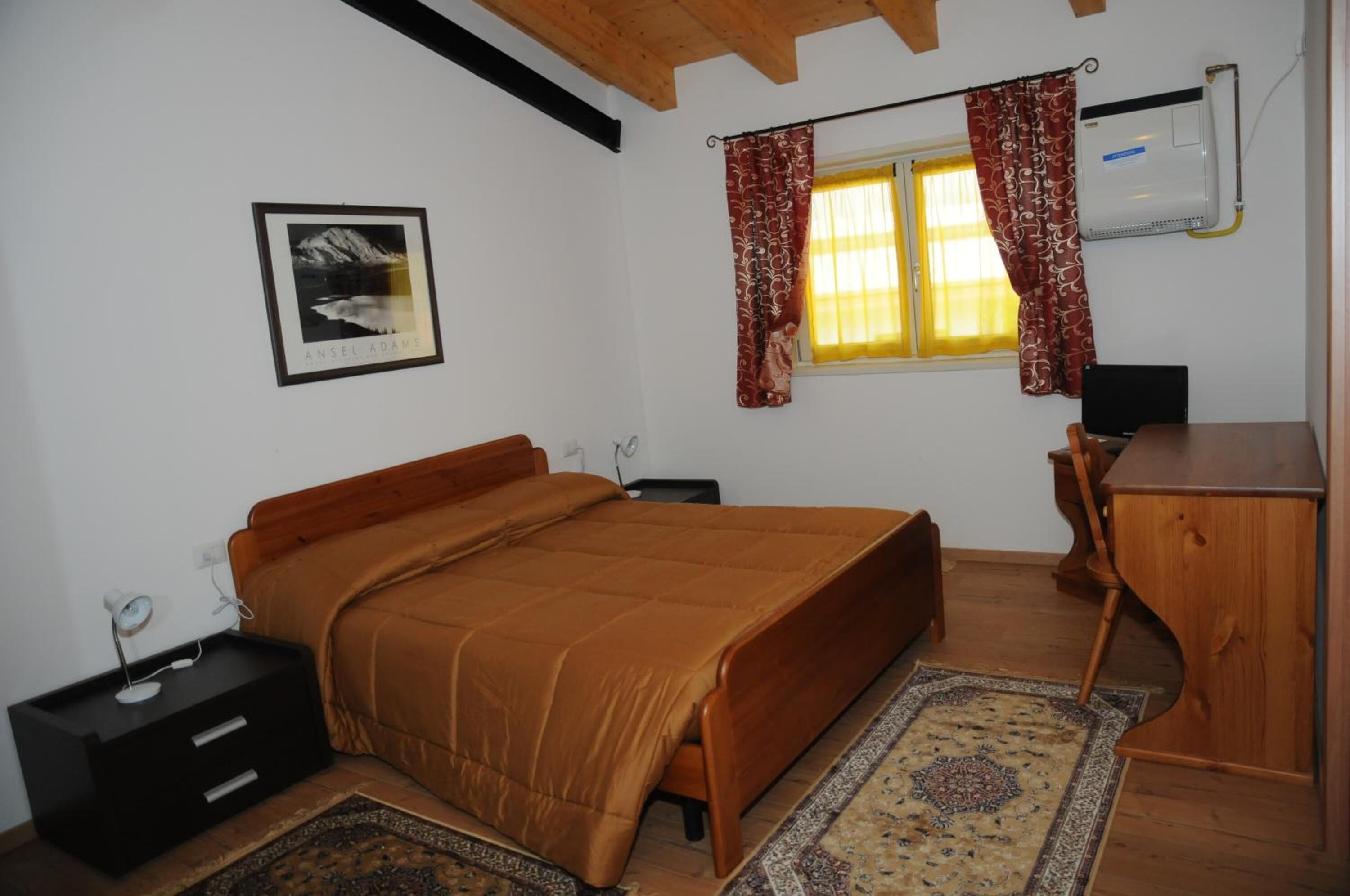 A bedroom with a bed, a desk, a chair, a small bedside table, a wall-mounted air conditioner, a window with curtains, and a framed picture on the wall.
