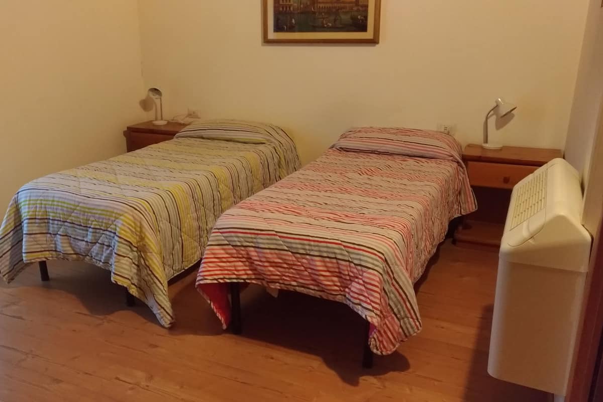 Quarto com duas camas de solteiro, piso de madeira, mesas de cabeceira com abajures e uma pequena escrivaninha com teclado.