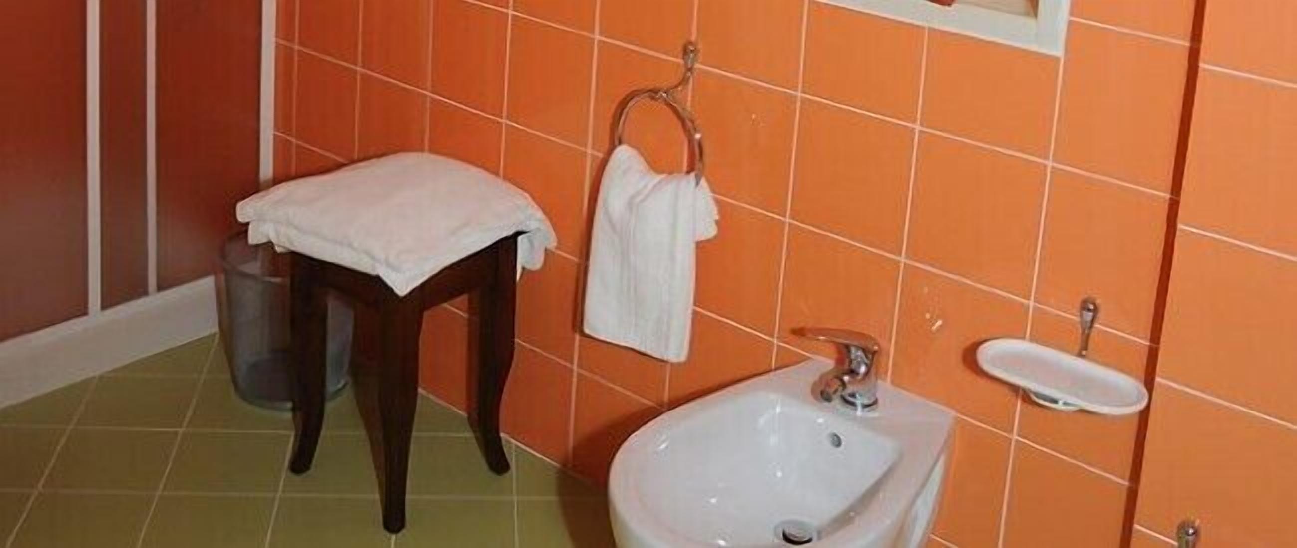 Een badkamer met een oranje betegelde muur, een witte wastafel, een houten kruk en een metalen prullenbak.