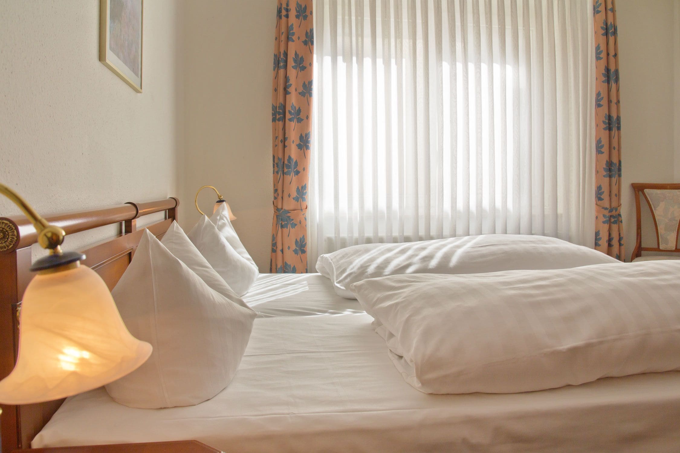 Un dormitorio con dos camas, una lámpara de noche y cortinas translúcidas con estampado floral.