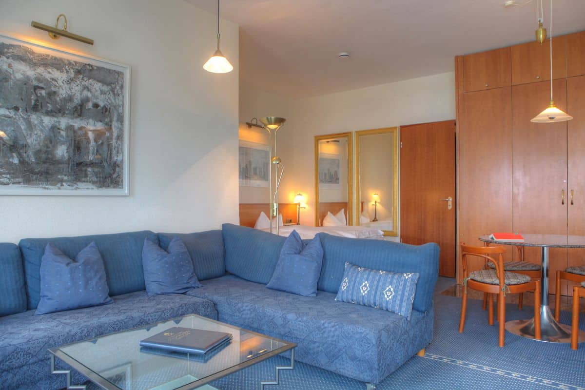 A modern living room with a blue sofa, a glass coffee table, and a dining area with a wooden table and chairs.