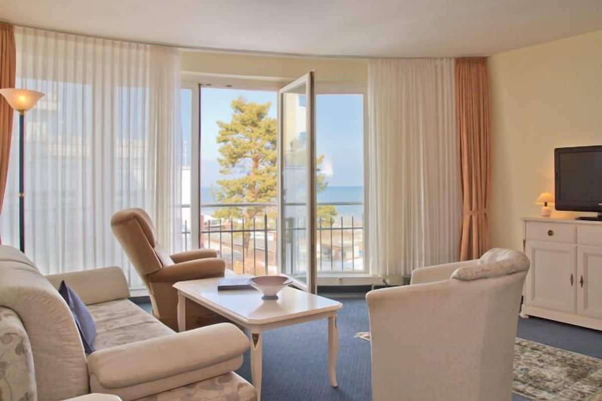 A hotel room with a sofa, armchair, and a small table with a bowl. There is a television on a cabinet and a view of the sea through large windows.