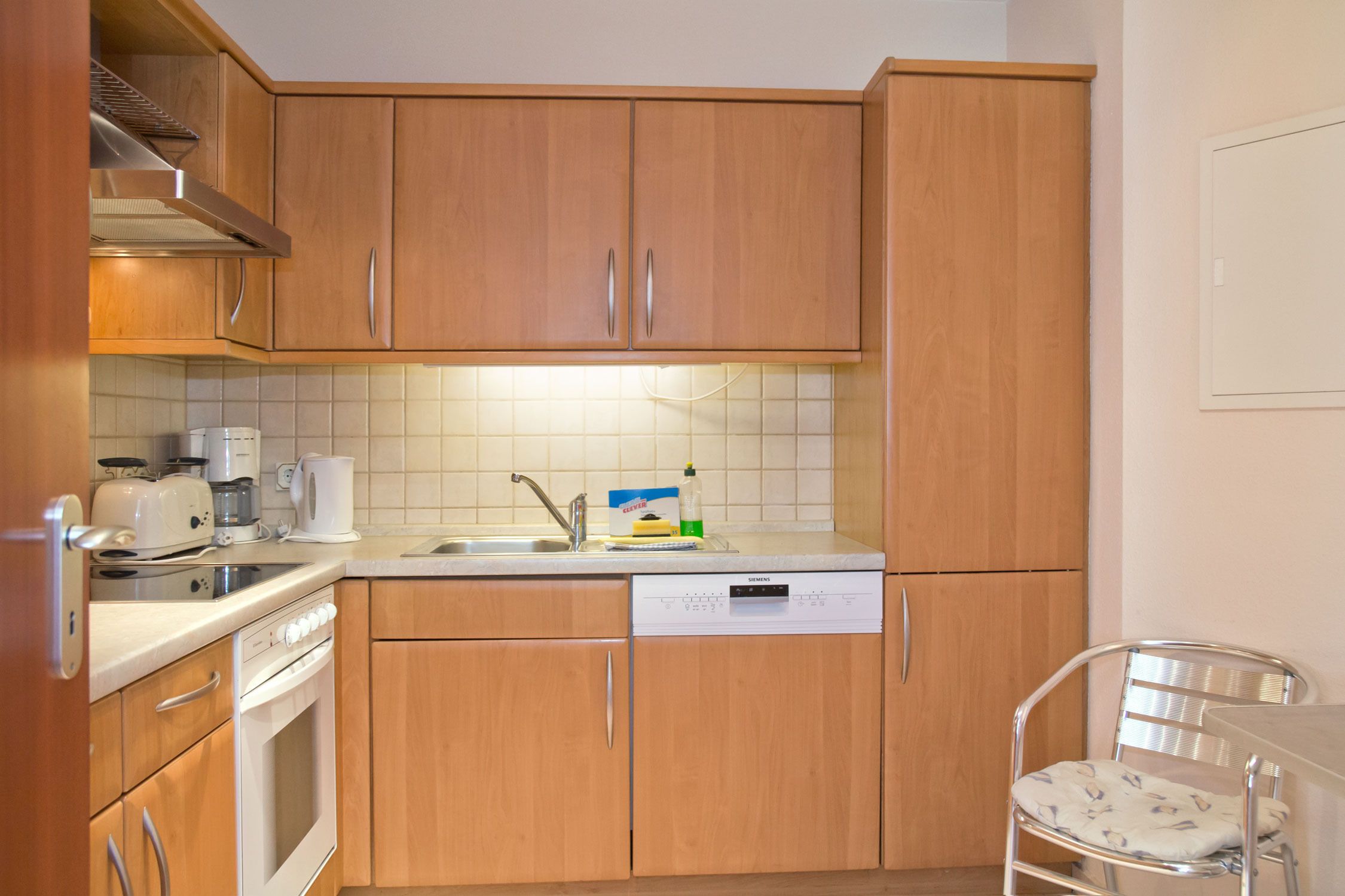 Una cocina compacta con armarios de madera, lavavajillas, estufa y un pequeño comedor con una silla.