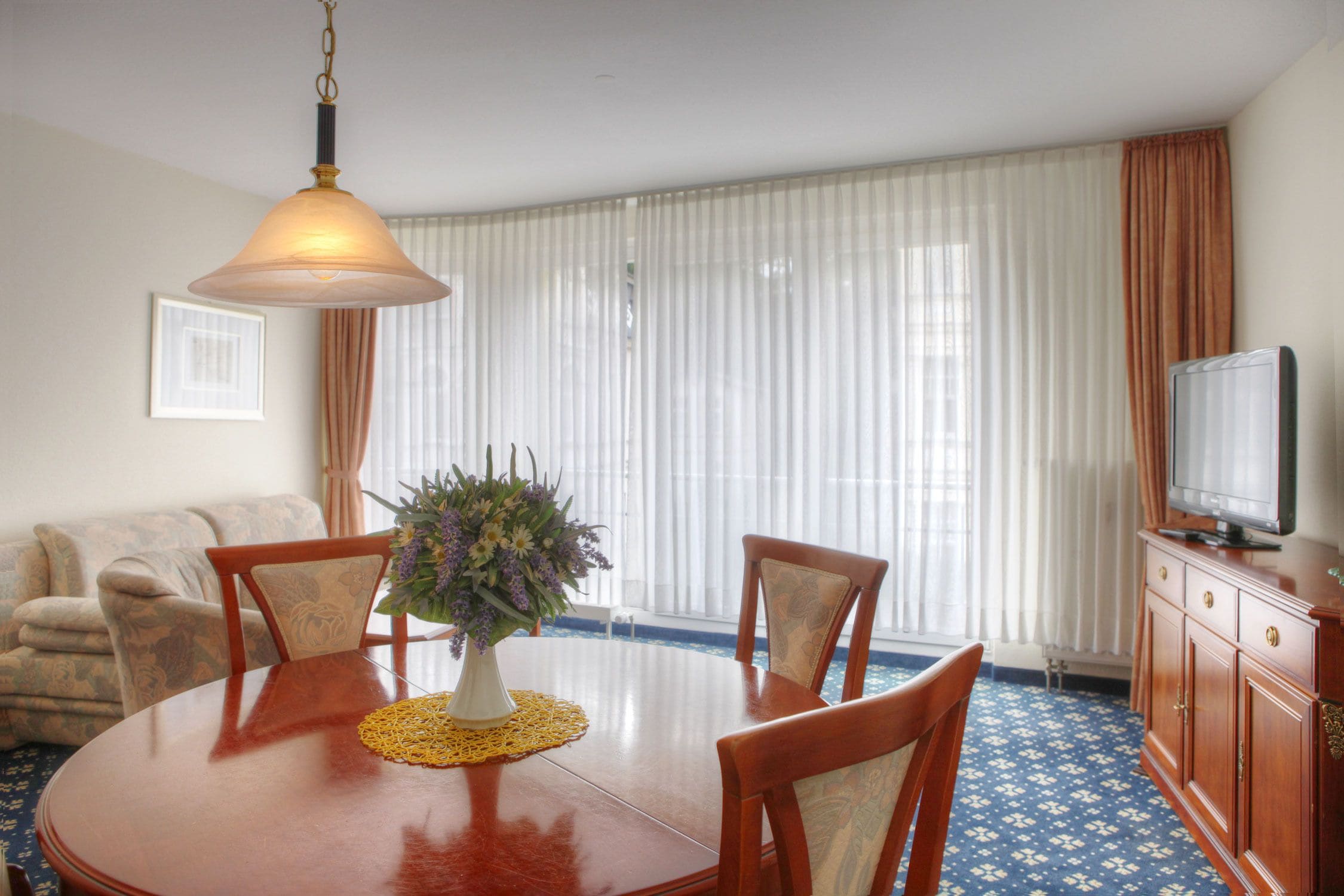 A dining area with a round table, chairs, a vase with flowers, a television on a cabinet, and a window with curtains.