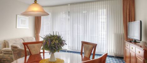 A dining area with a round table, chairs, a vase with flowers, a television on a cabinet, and a window with curtains.