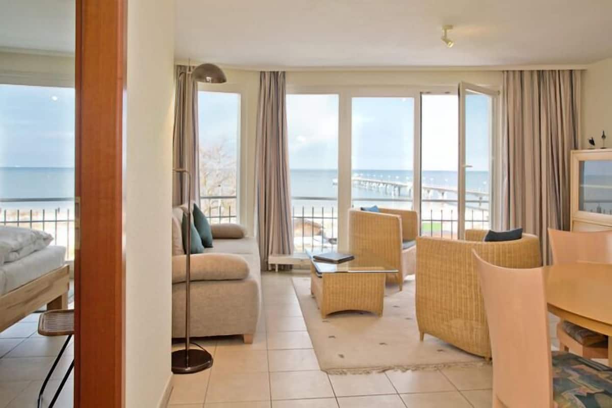 A hotel room with a large window offering a view of the sea and a bridge. There is a sofa, a chair, and a small table.
