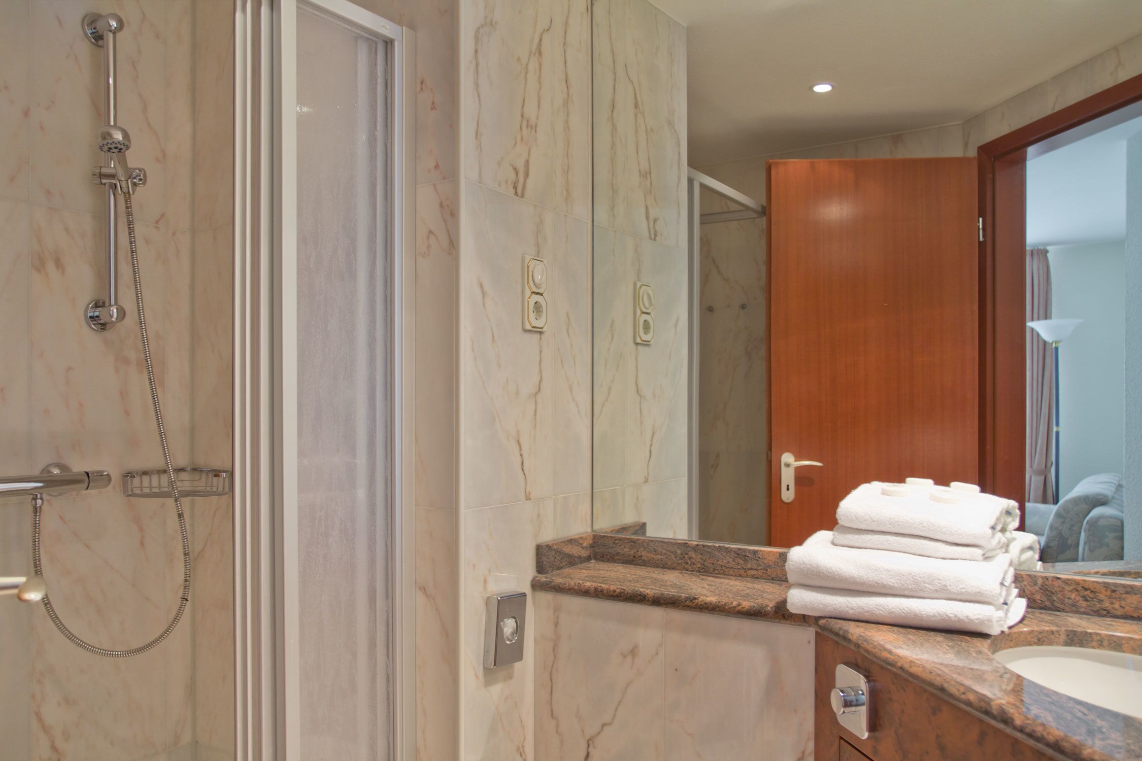 Un baño con pared de mármol, ducha, lavamanos con encimera de mármol y puerta de madera.