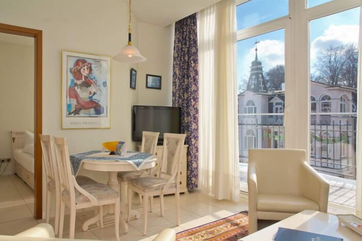 A room with a dining table, chairs, a television, and a large window with a view of buildings.