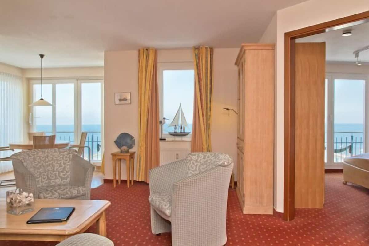A hotel room with a view of the sea, a dining area with a table and chairs, and a cozy seating area with patterned chairs.