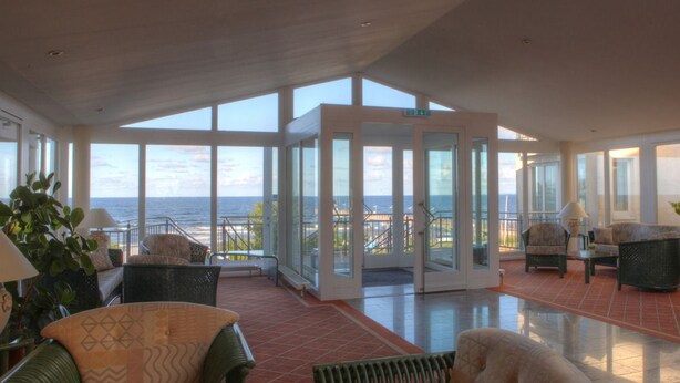 A spacious indoor area with large windows offering views of the sea, furnished with chairs and a table, and a glass door leading outside.