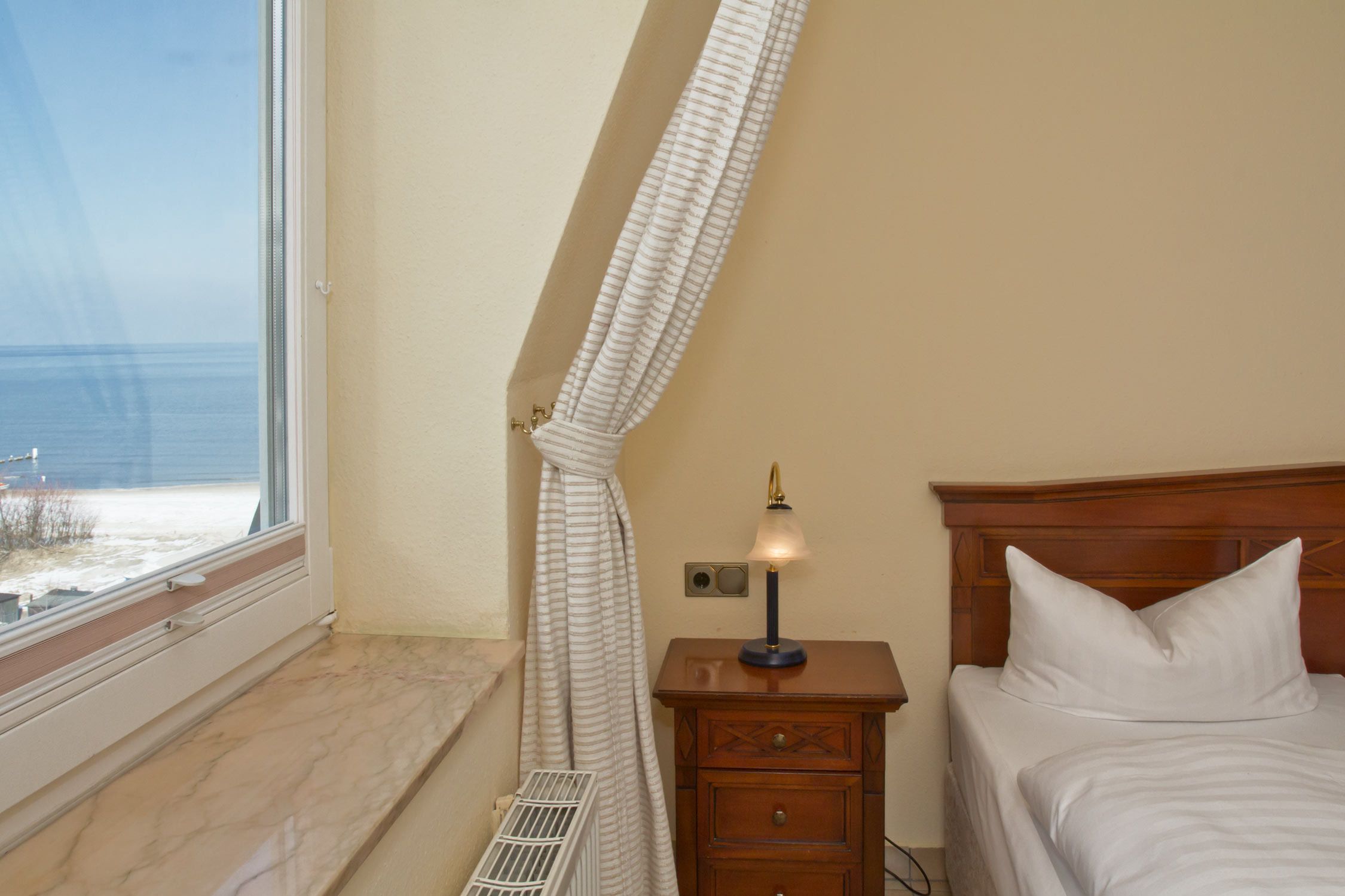 Une chambre à coucher avec un lit, une table de chevet avec une lampe, une fenêtre donnant sur la mer et un radiateur.