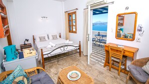 A room with a bed, a desk, a wicker chair, a small table, and a view of the sea through an open door. - Stelios Rooms (Santorini)