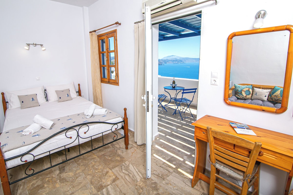 A bedroom with a bed, a desk, and a chair. There is a window with curtains and a view of the sea.