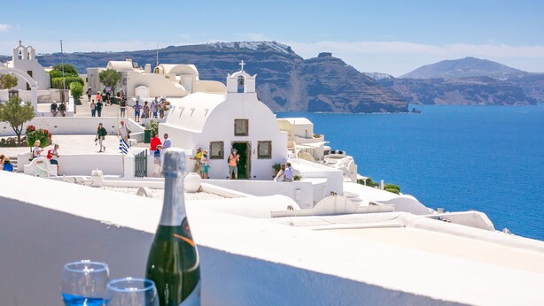 A coastal view with white buildings, a bottle of wine, and two glasses on a table. - Stelios Rooms (Santorini)