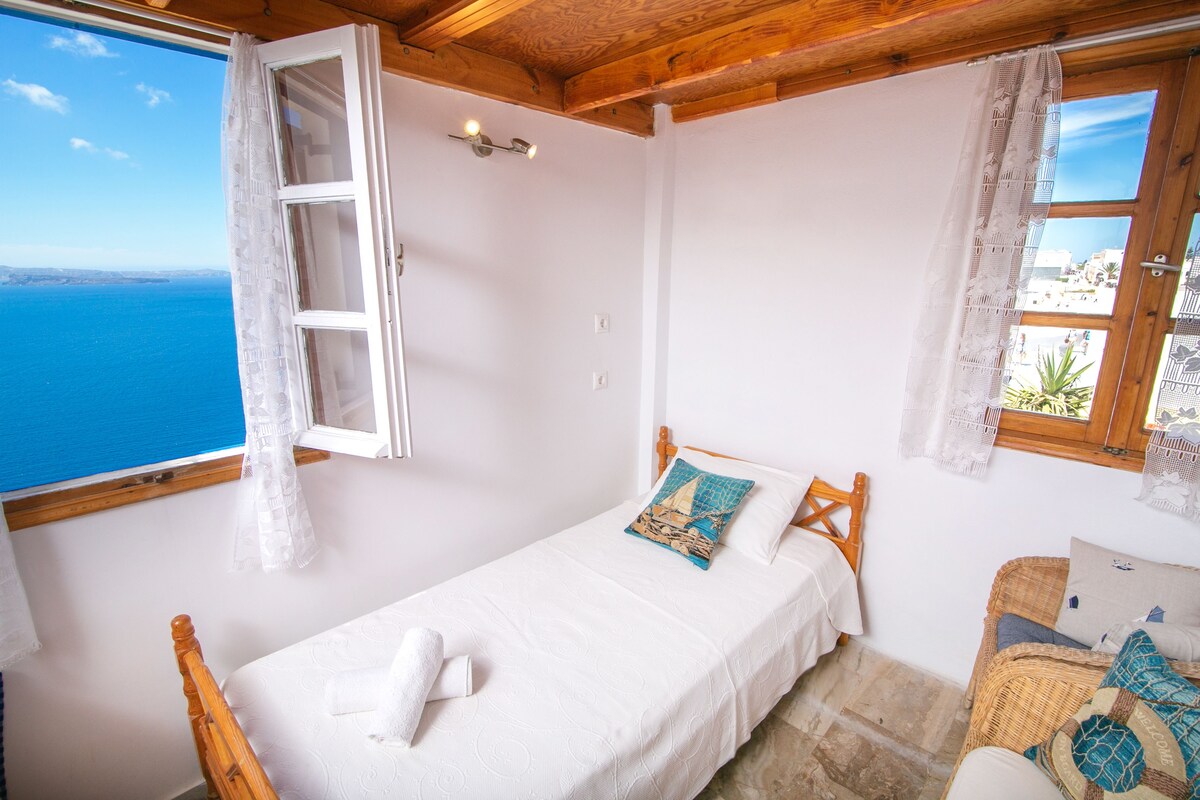 A bedroom with a bed, a wooden headboard, a window with a view of the sea, and a small balcony with a wicker chair.