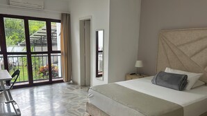 Ein Schlafzimmer mit einem Bett, einem Nachttisch, einer Lampe, einem Sessel und Blick auf einen Balkon mit Pflanzen.