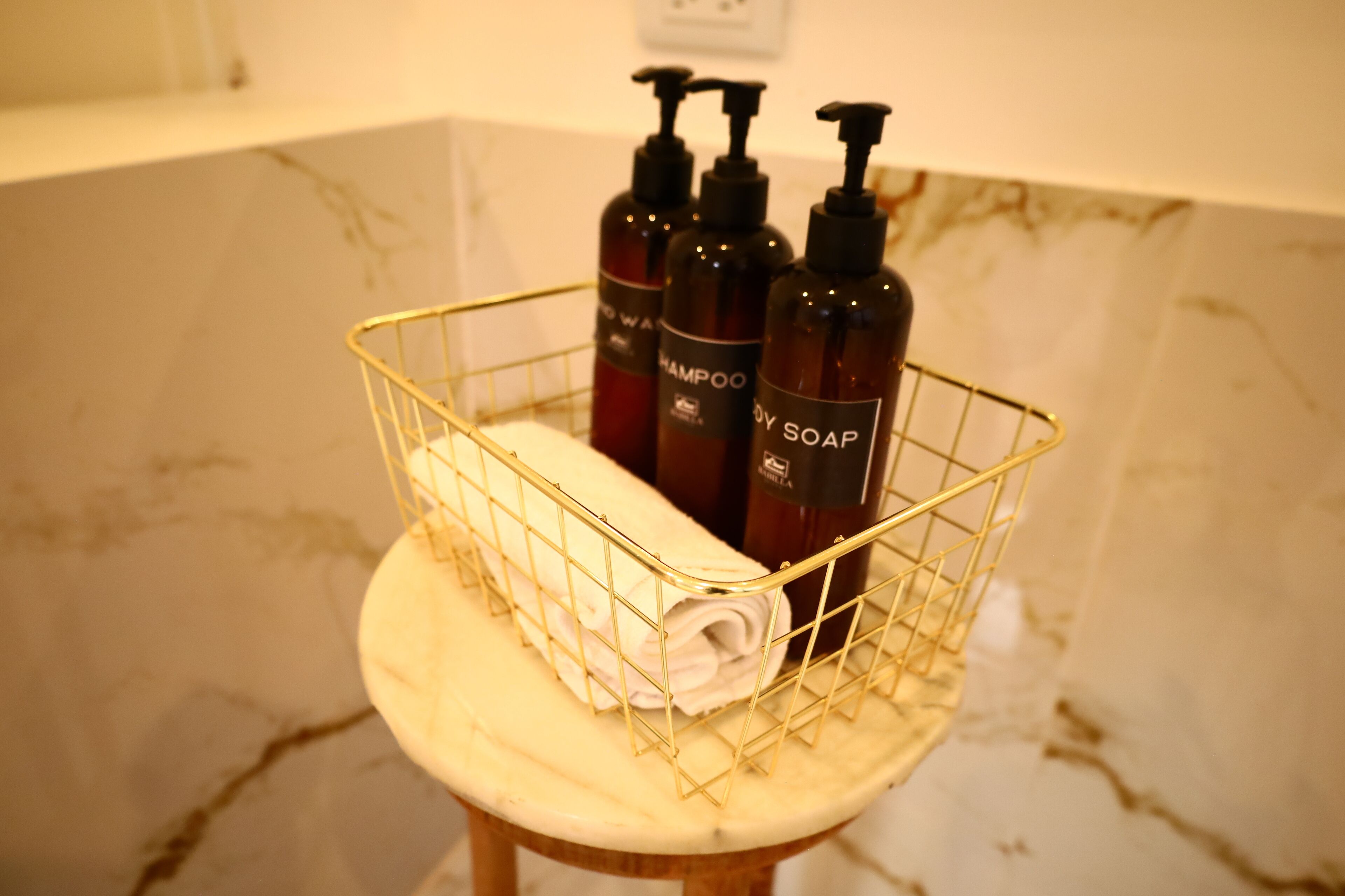 A bathroom with a marble countertop, a gold wire basket holding shampoo and body soap bottles, and a rolled towel.