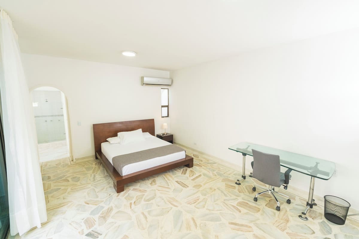 A bedroom with a bed, a desk, a chair, an air conditioning unit, and a shower area.