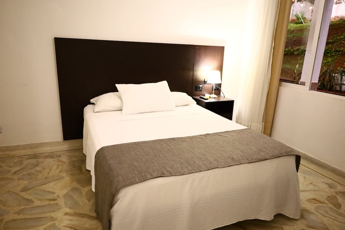 A neatly made bed with a headboard, a bedside lamp, and a window with a view of greenery.