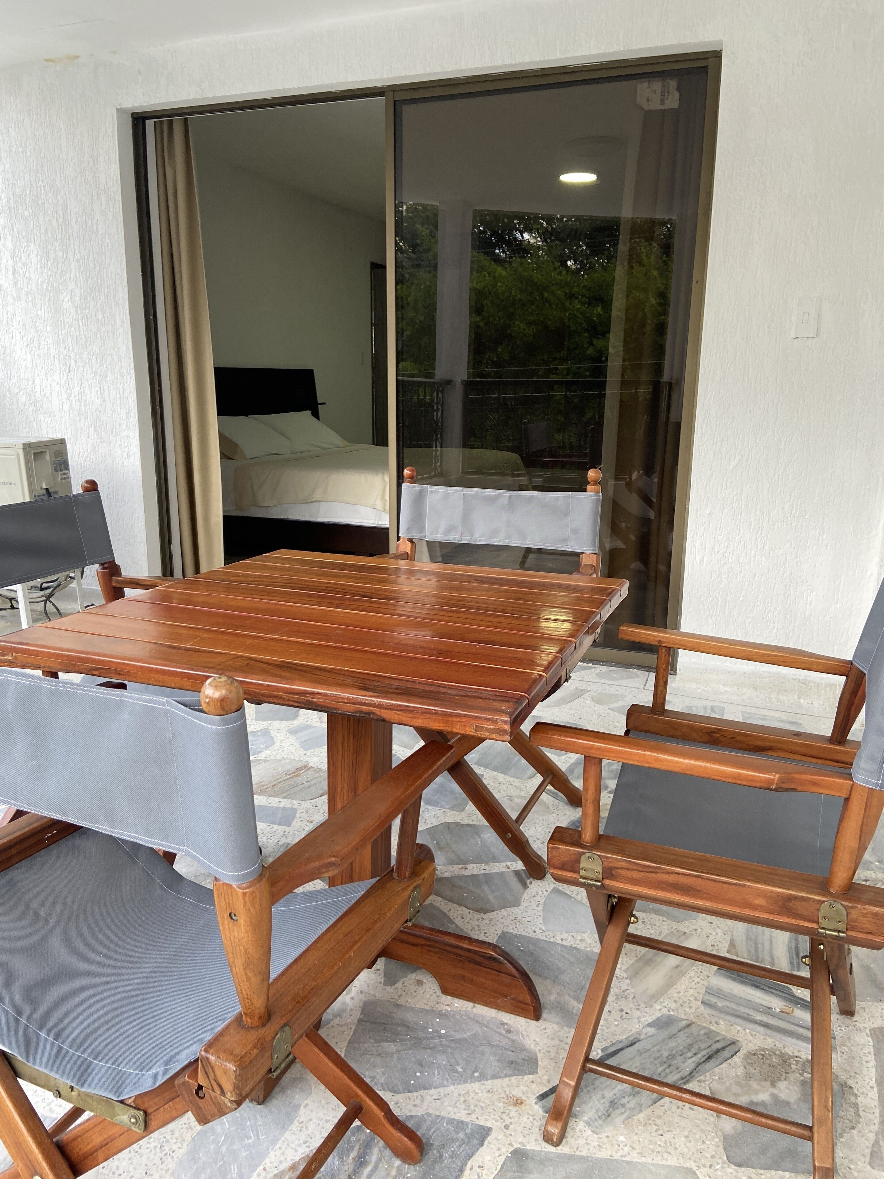 Una mesa de comedor de madera con sillas acolchadas, una cama con cabecera blanca y un ventanal corredero que da a un espacio exterior.