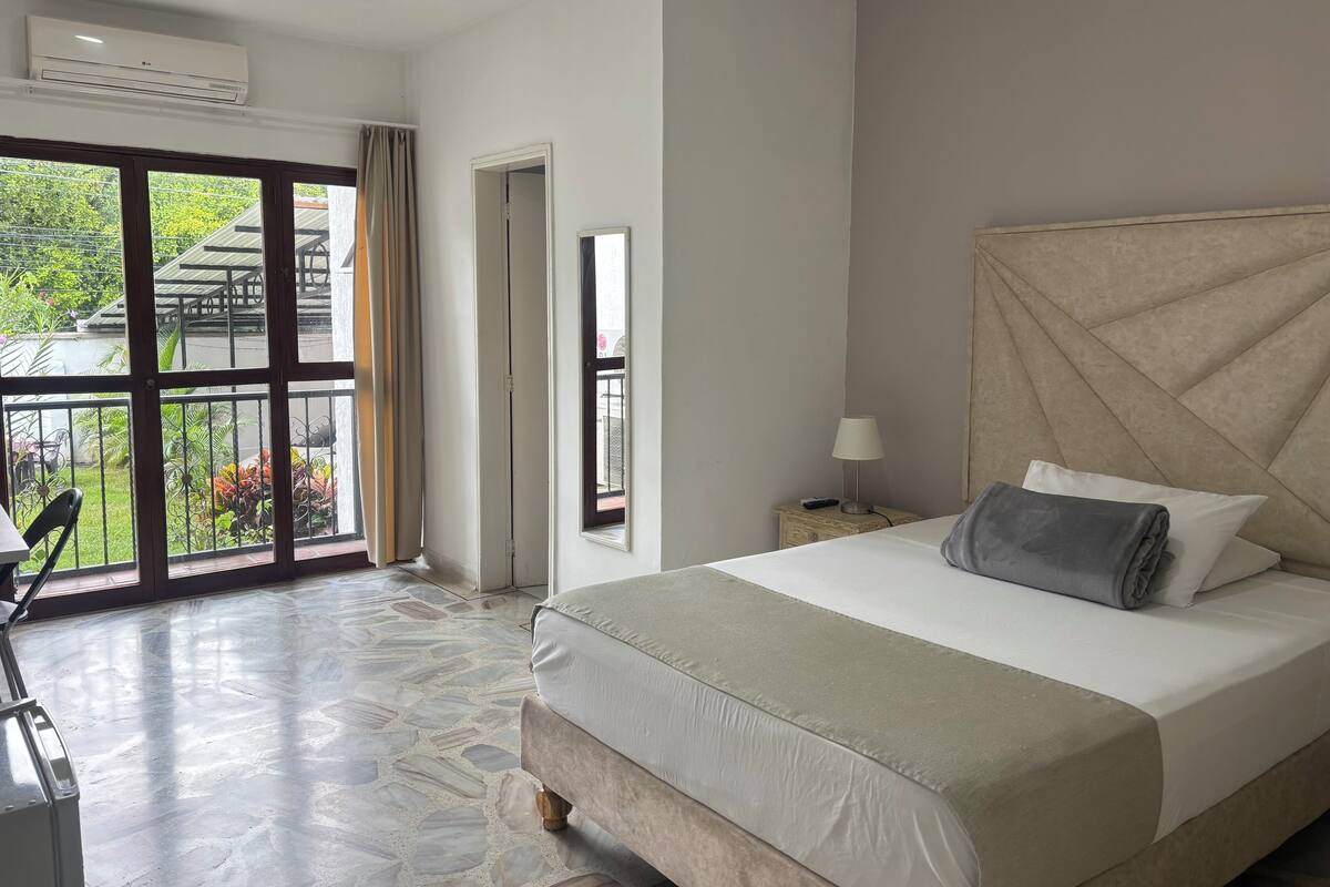 A hotel room with a large bed, a chair, a desk, and a balcony with a view of greenery and a building.