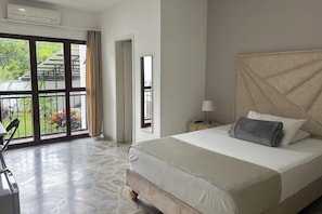 Quarto de hotel com uma cama grande, uma cadeira, uma mesa e varanda com vista para vegetação e um edifício.