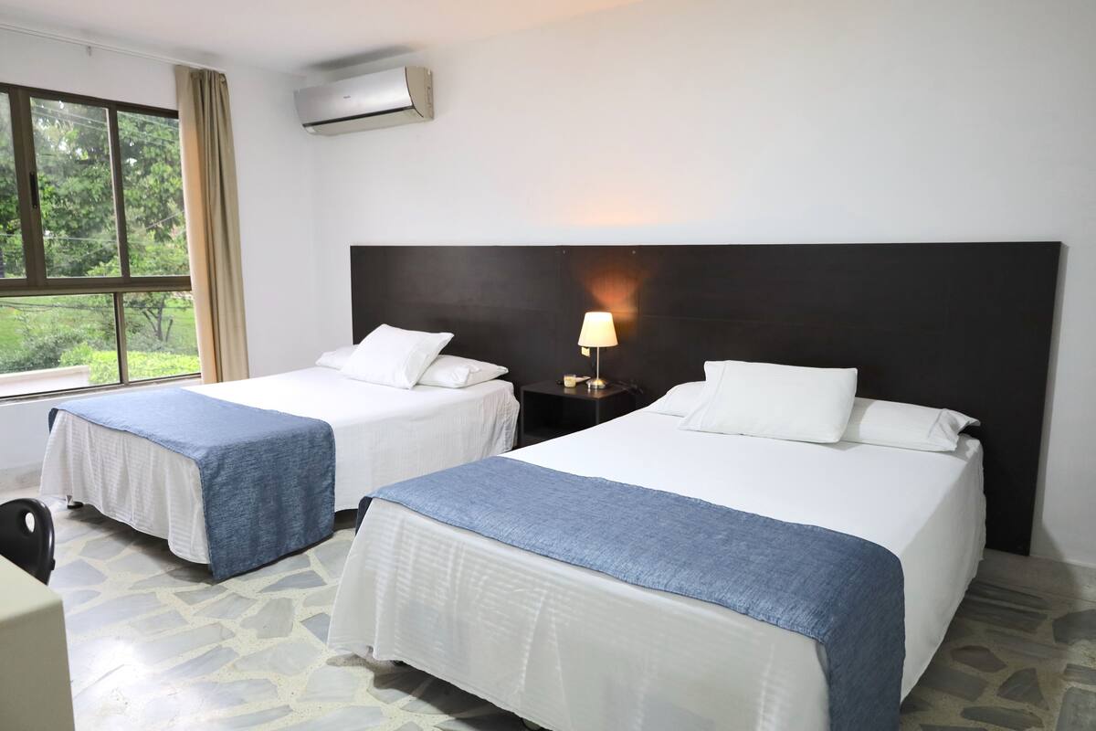A hotel room with two beds, a nightstand with a lamp, a window with curtains, and an air conditioning unit.
