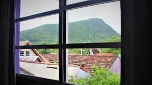 A window view of a mountain with a red-roofed building in the foreground. - Pousada Santa Ana (Florianópolis)