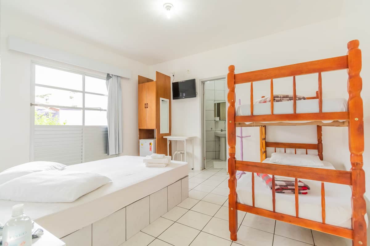 A hotel room with a bunk bed, a single bed, a desk with a chair, a television, and a bathroom visible through an open door.