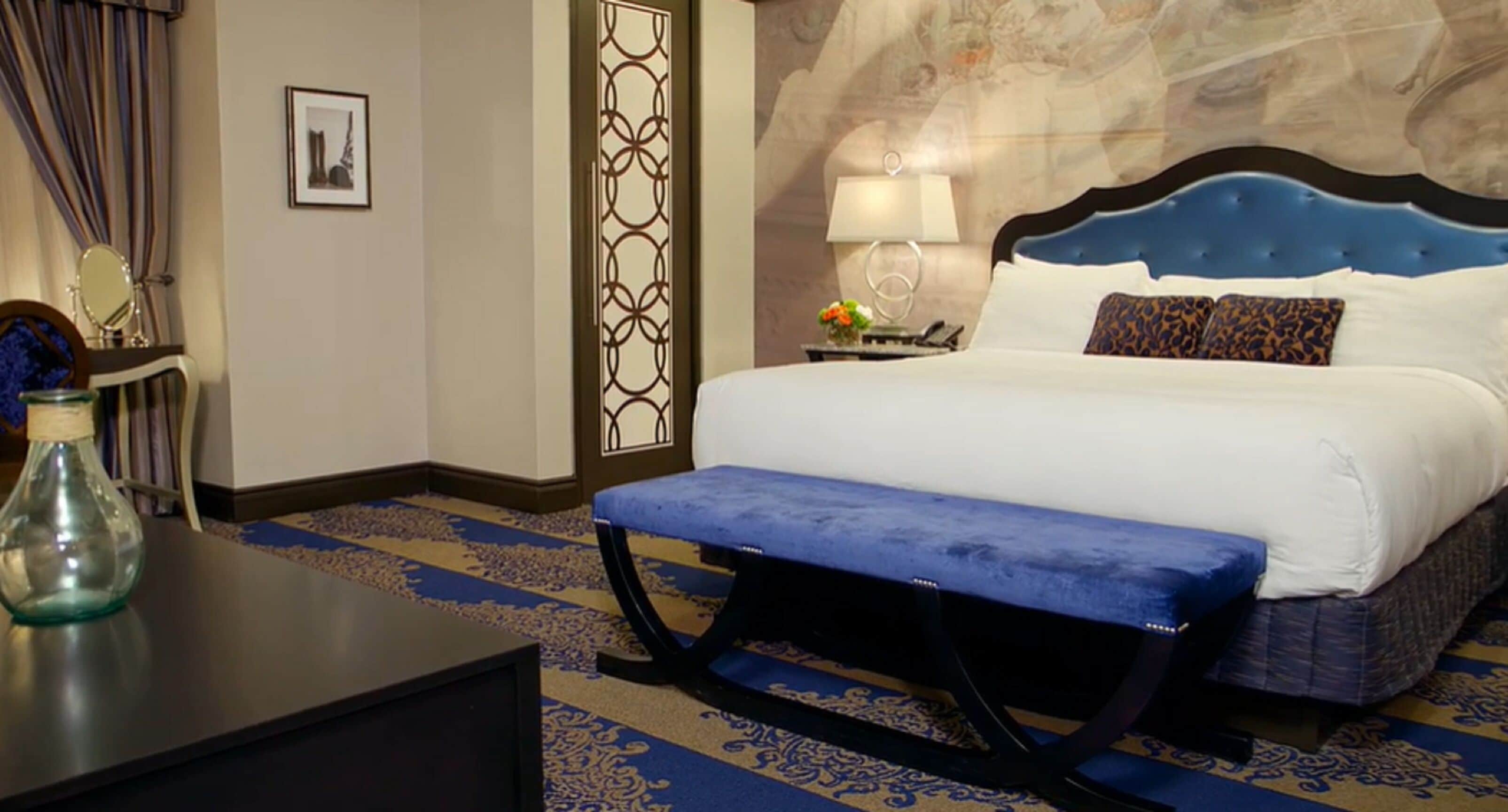 Une chambre d’hôtel comprenant un lit, une banquette, une table de chevet, une lampe, un miroir et un panneau décoratif mural.