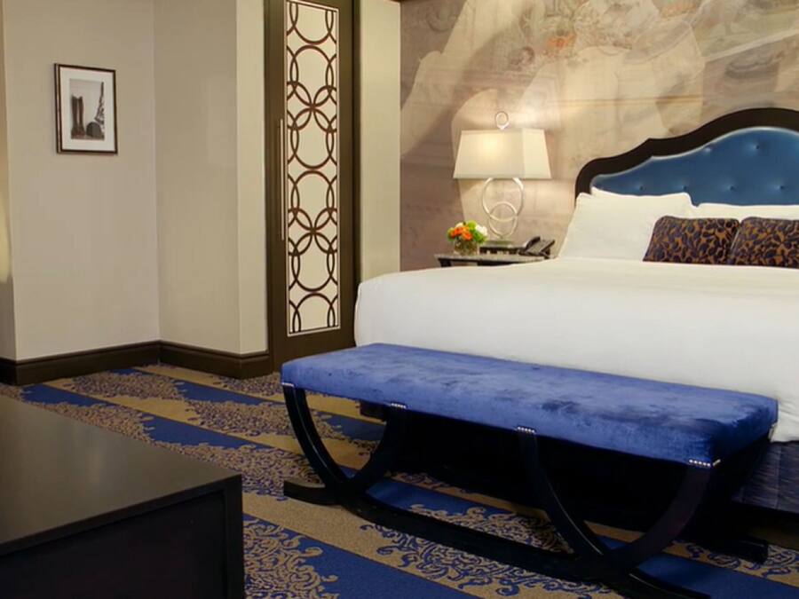 A hotel room with a bed, a bench, a nightstand, a lamp, a mirror, and a decorative wall panel.