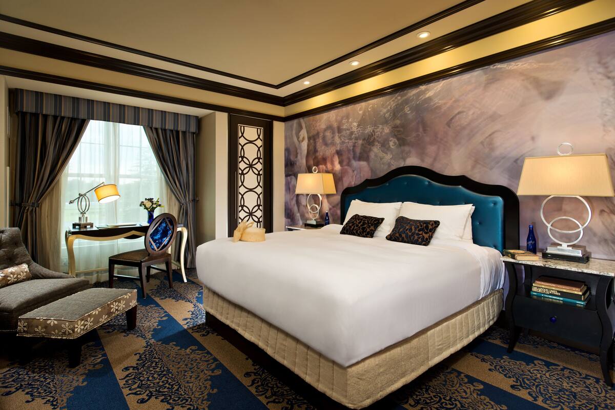 Une chambre d’hôtel spacieuse avec un grand lit, un coin salon comprenant un fauteuil et un pouf, un bureau avec une lampe et un mur décoratif.