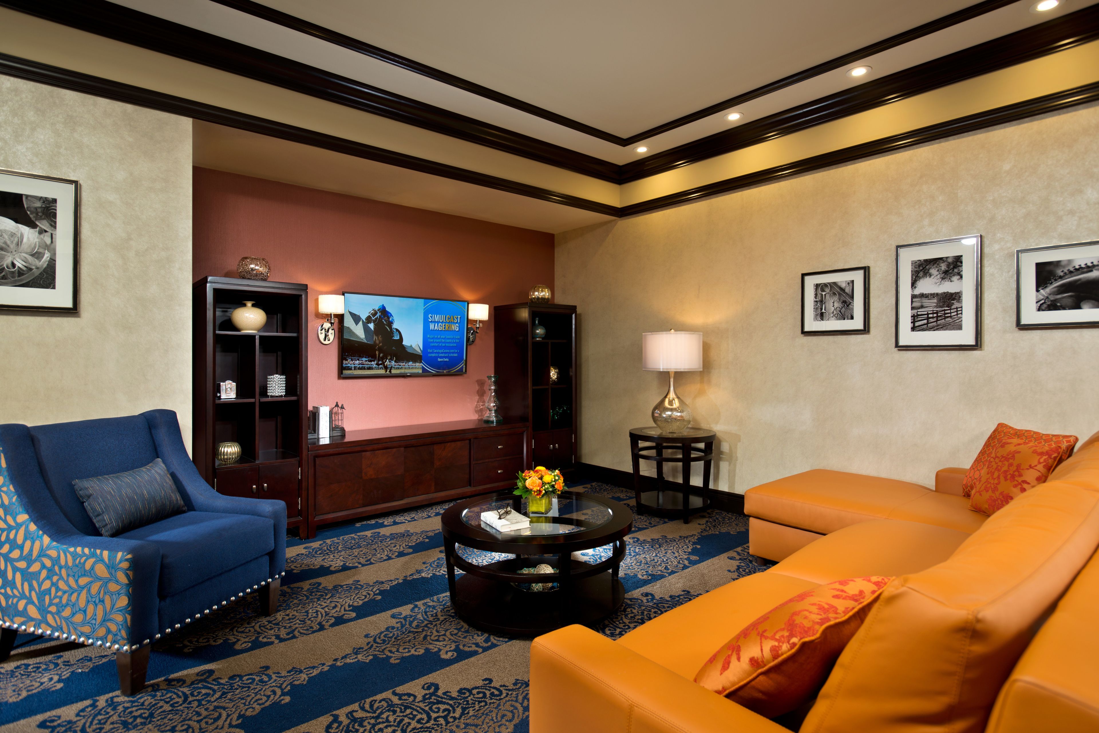 A hotel lobby with a blue armchair, an orange sofa, a round coffee table, a television, and framed pictures on the wall.