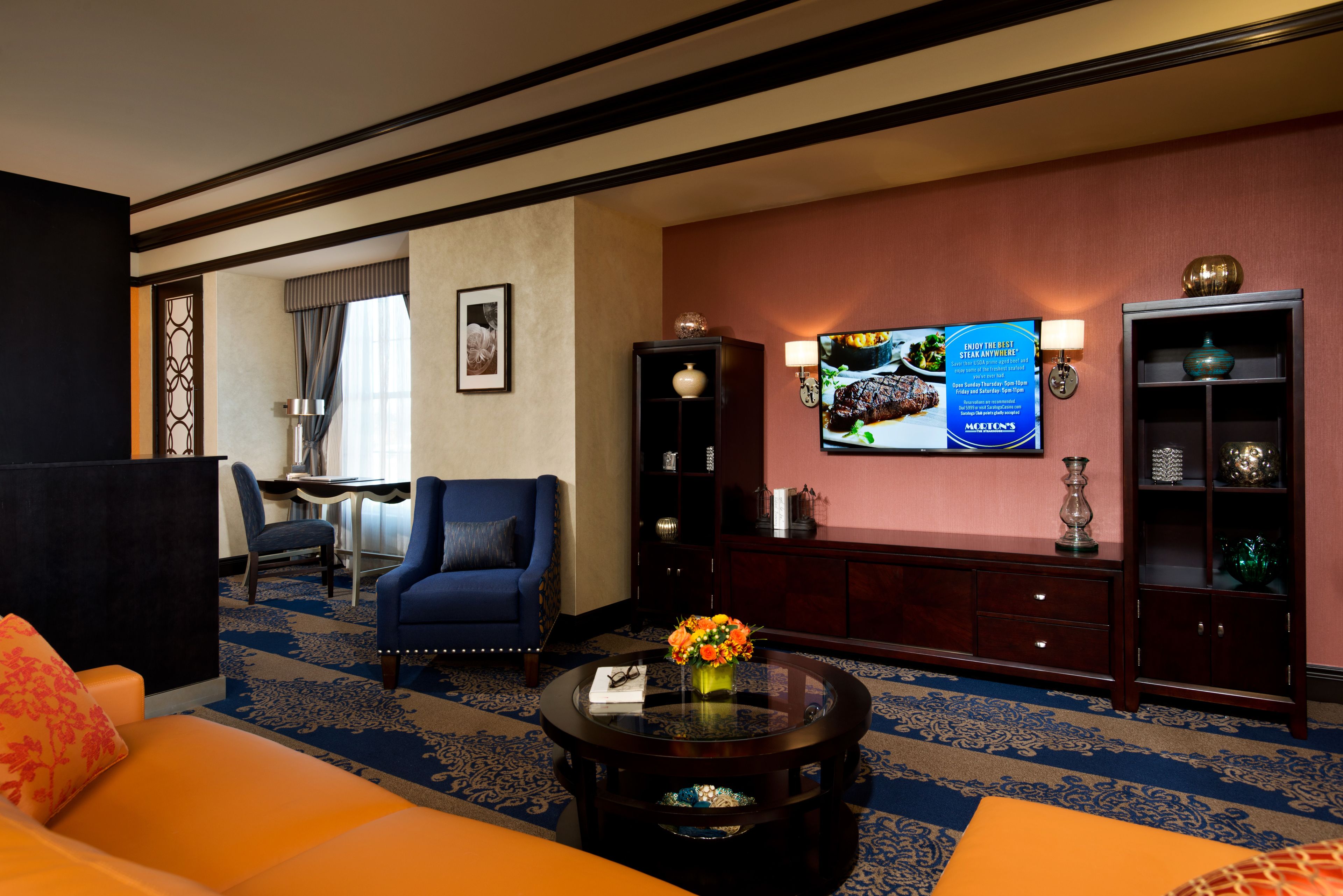 Una habitación de hotel con un sillón azul, un sofá, una mesa de centro con un florero, un televisor y un mueble con adornos.