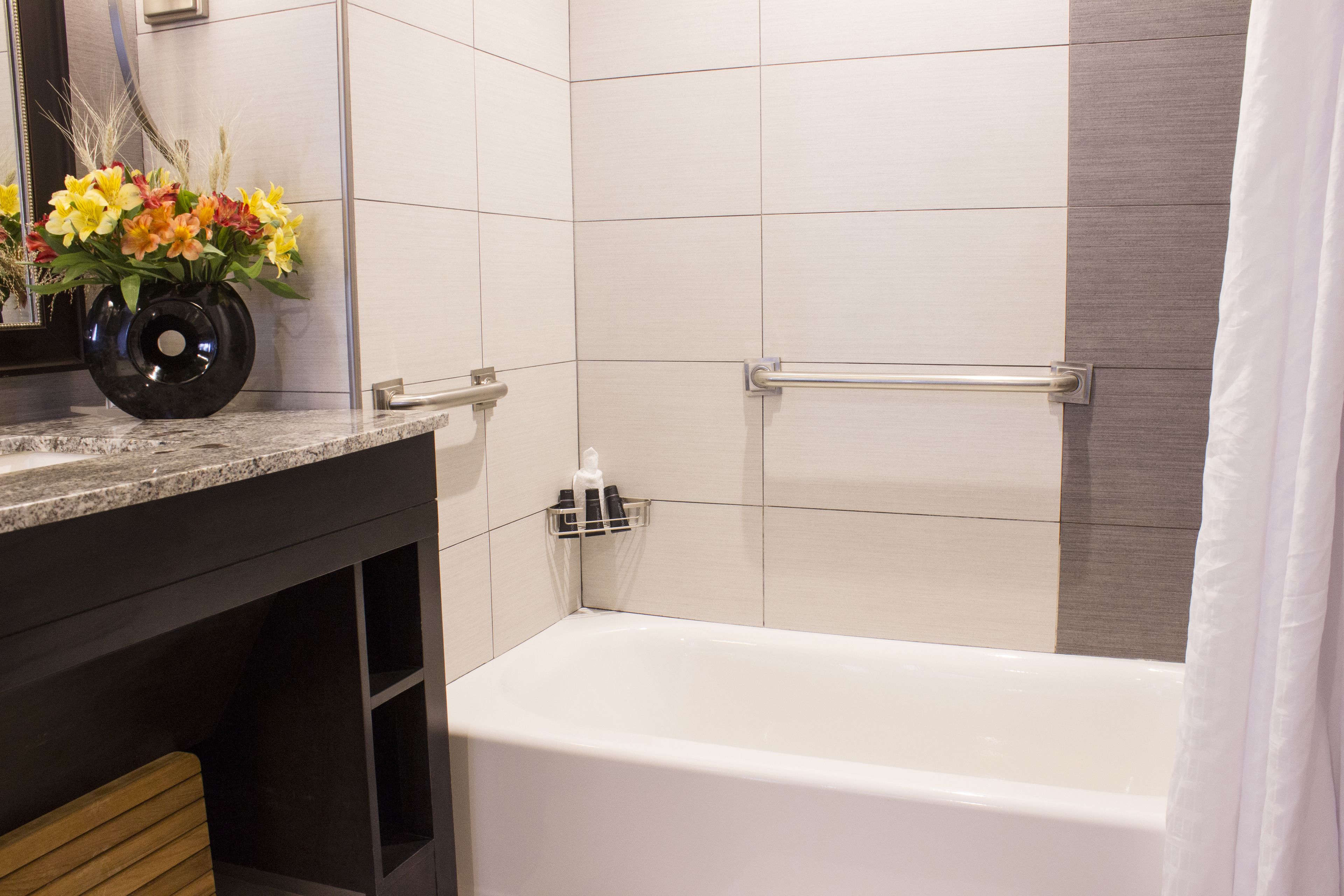 A bathroom with a white bathtub, a vanity with a black vase of flowers, and a handrail.