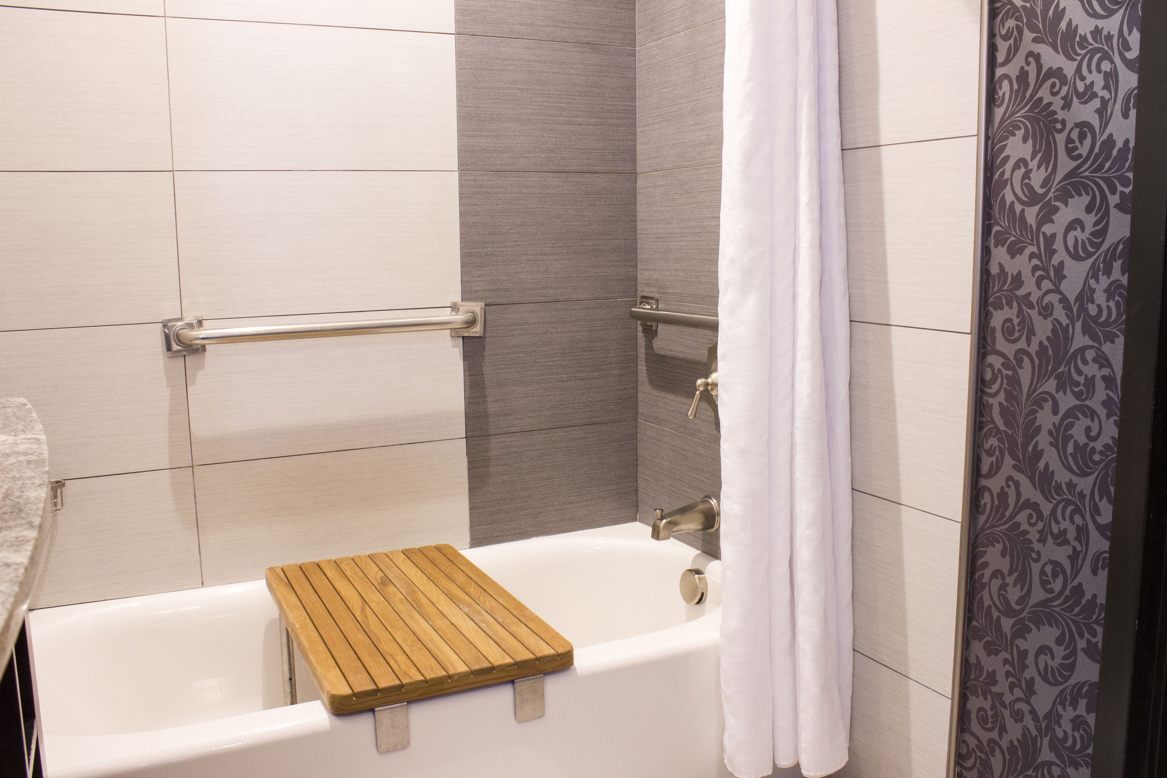 A bathroom with a bathtub, wooden bath mat, grab bars, and a shower curtain.