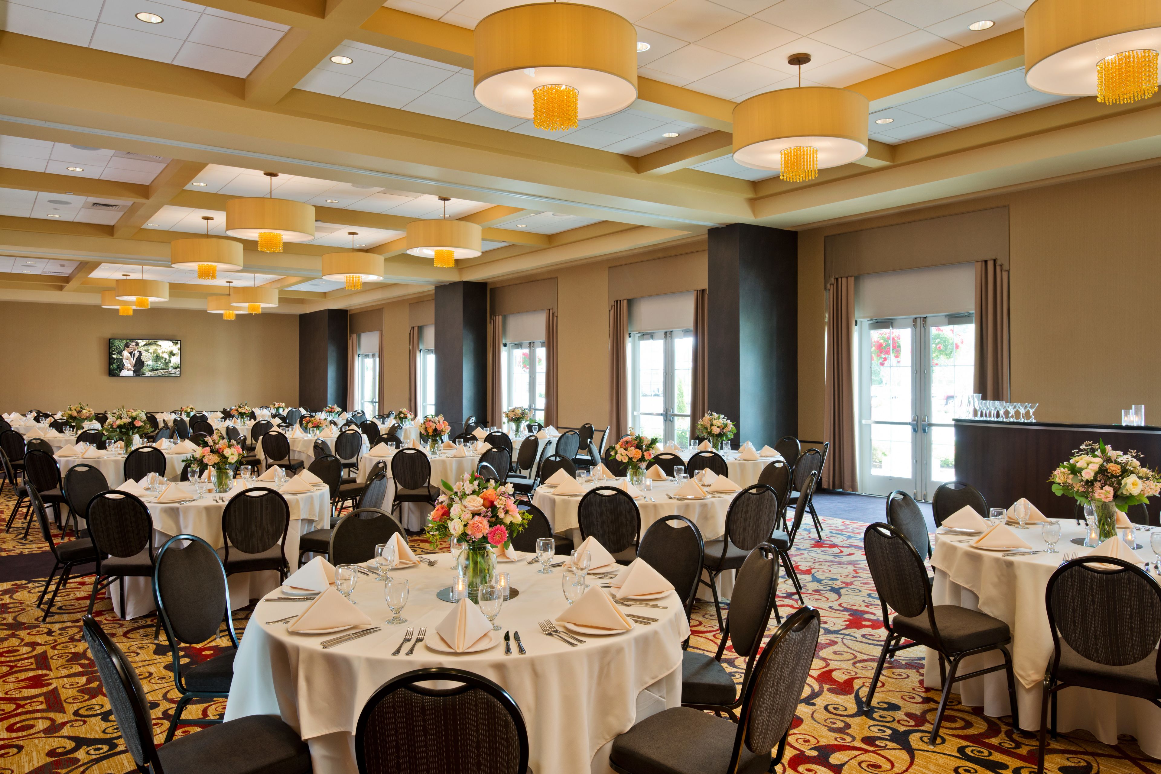Un salón de banquetes con mesas redondas dispuestas para el servicio de comidas, equipado con sillas, vajilla y centros de mesa florales.