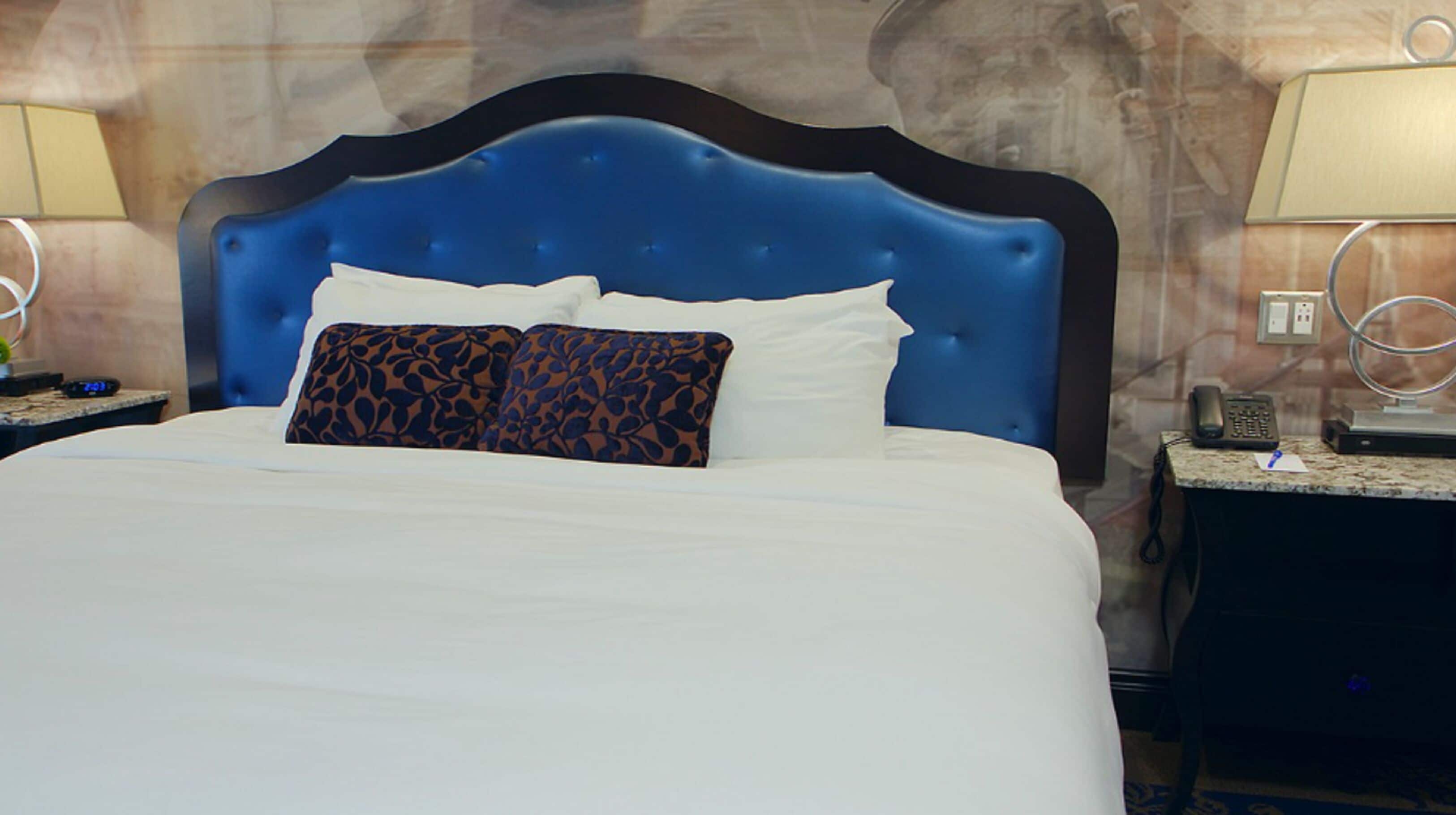 A hotel room with a large bed, a blue headboard, white bedding, and a nightstand with a phone and lamp.
