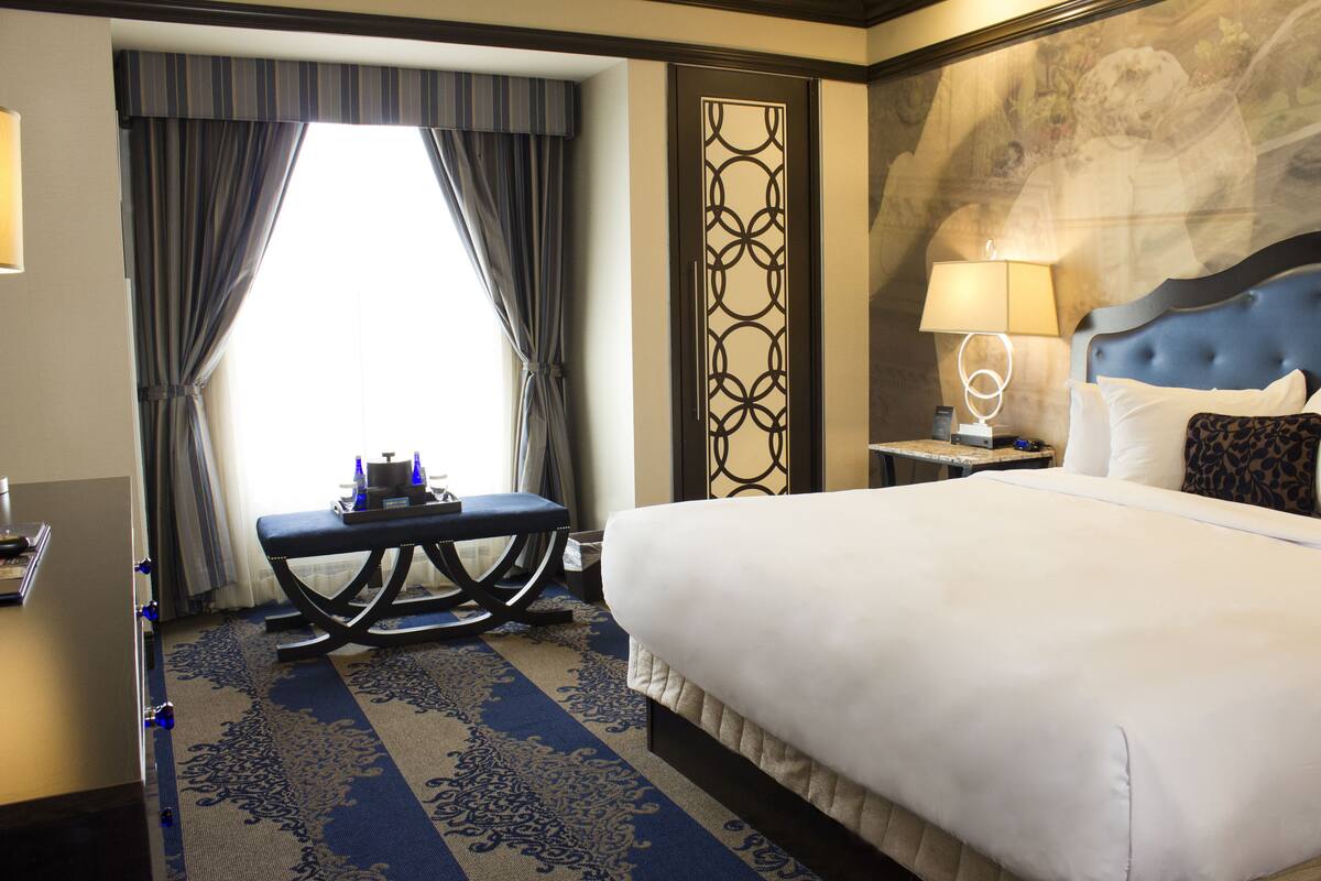 Une chambre d’hôtel avec un grand lit, un bureau avec une chaise, une table de chevet avec une lampe et une vue sur l’extérieur par une fenêtre munie de rideaux.
