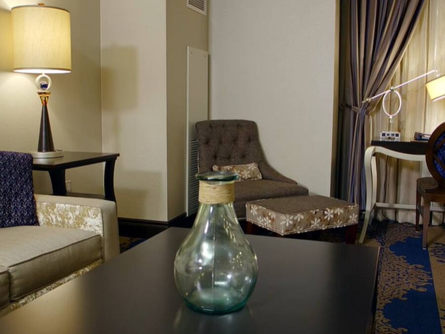 Habitación de hotel con sofá, una silla, una mesita con un florero, lámpara, espejo y cama con cortinas.