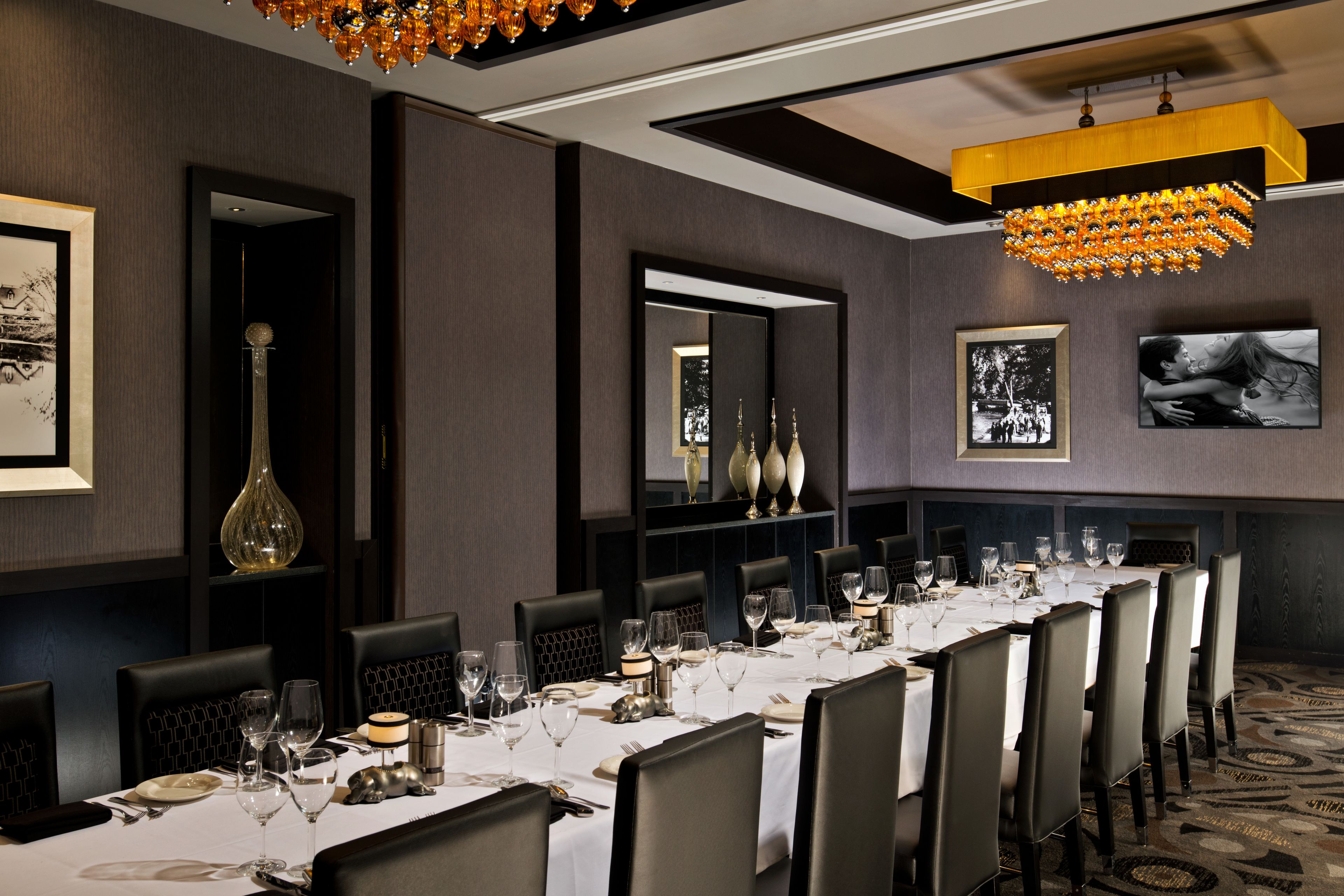 Una larga mesa de comedor dispuesta con manteles blancos, sillas y vajilla en un interior moderno con paredes oscuras, una araña y cuadros enmarcados.