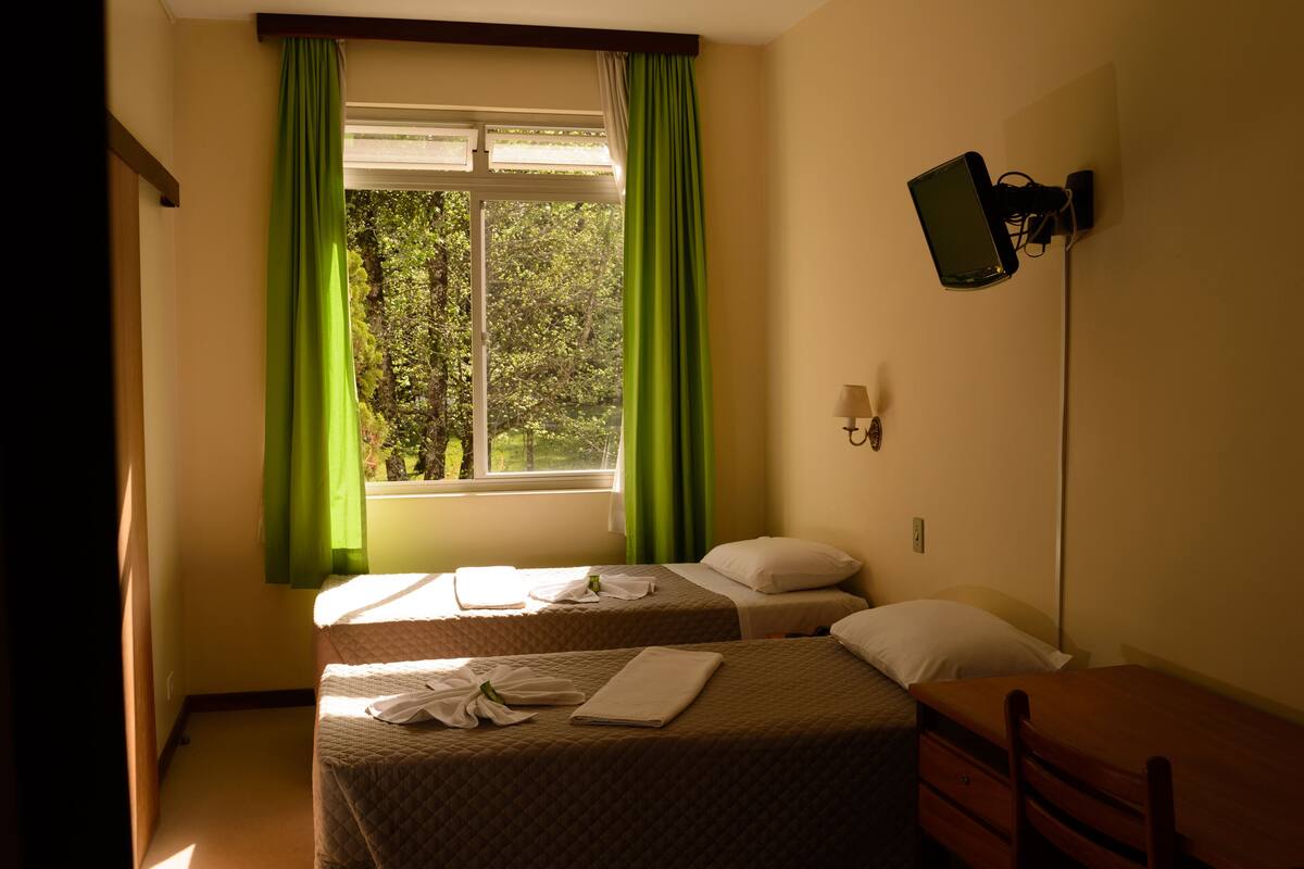 A hotel room with two beds, a desk, a chair, a TV mounted on the wall, and a window with green curtains.