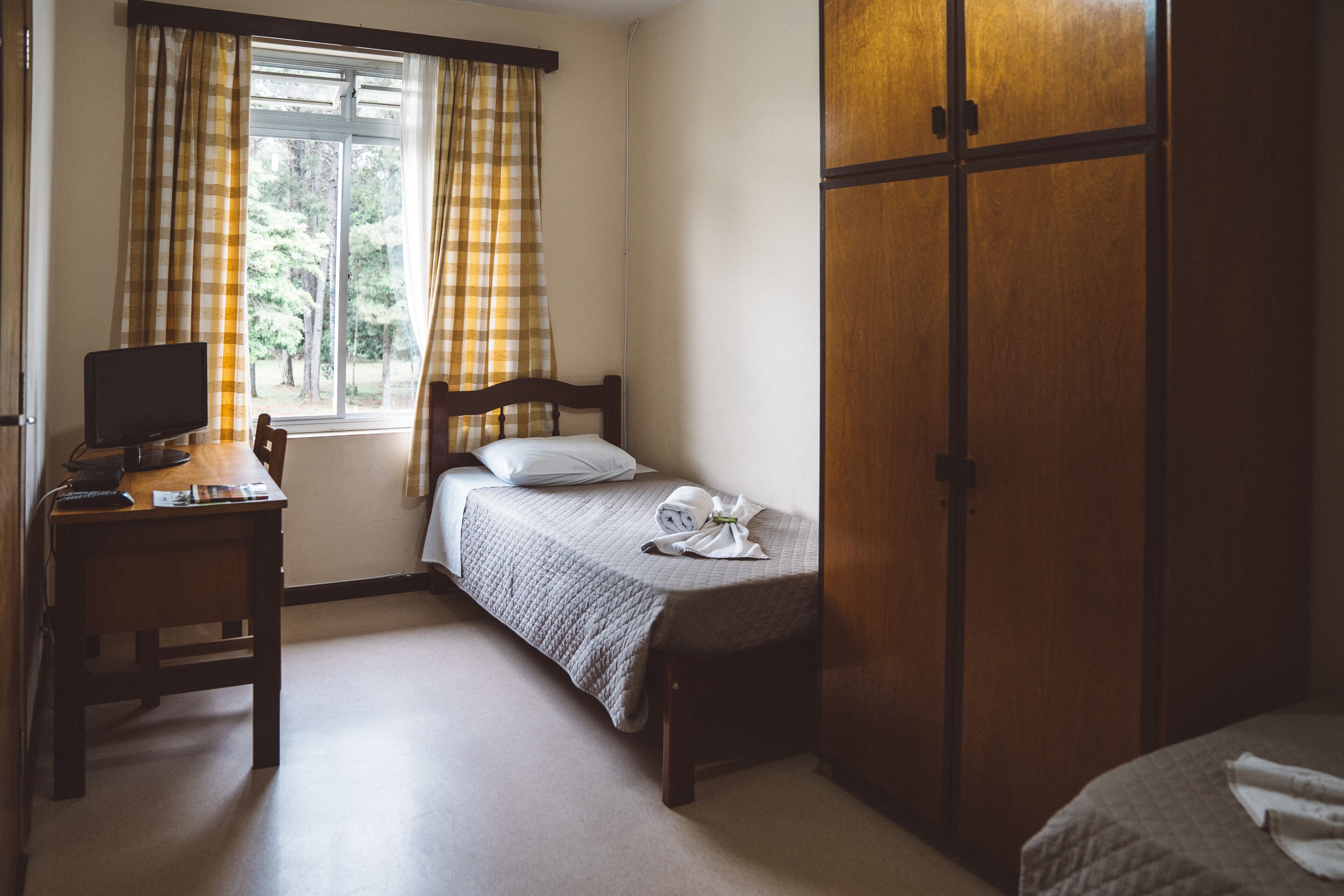 Un dormitorio con cama, escritorio con computadora, armario y ventana con cortinas.