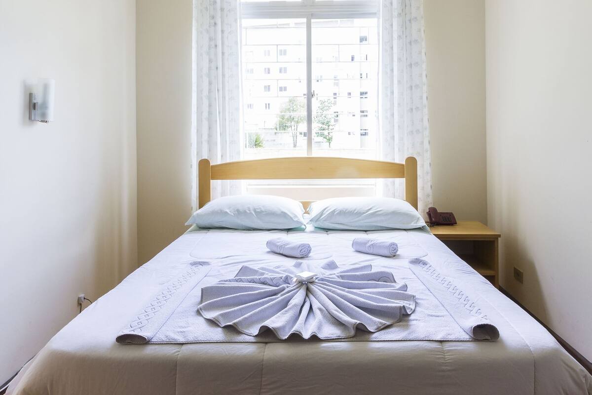 A single bed with a wooden frame and white bedding, a nightstand with a lamp, and a window with curtains.