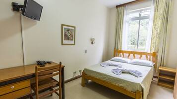 A bedroom with a wooden bed, a desk with a chair, a television mounted on the wall, and a window with curtains.
