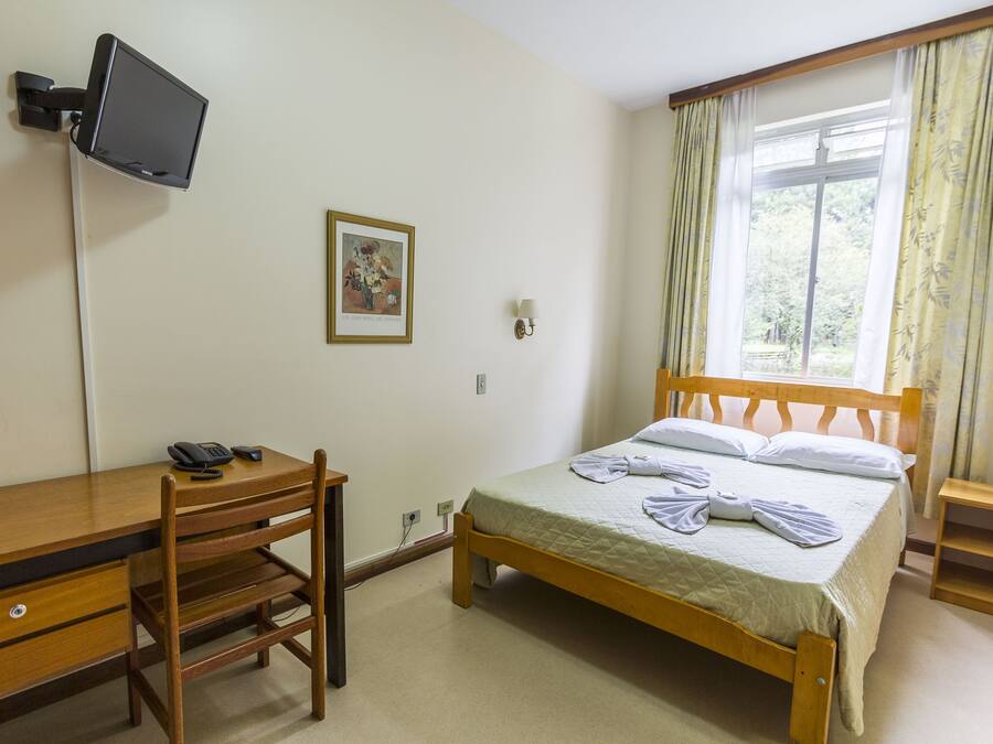 A bedroom with a wooden bed, a desk with a chair, a television mounted on the wall, and a window with curtains.