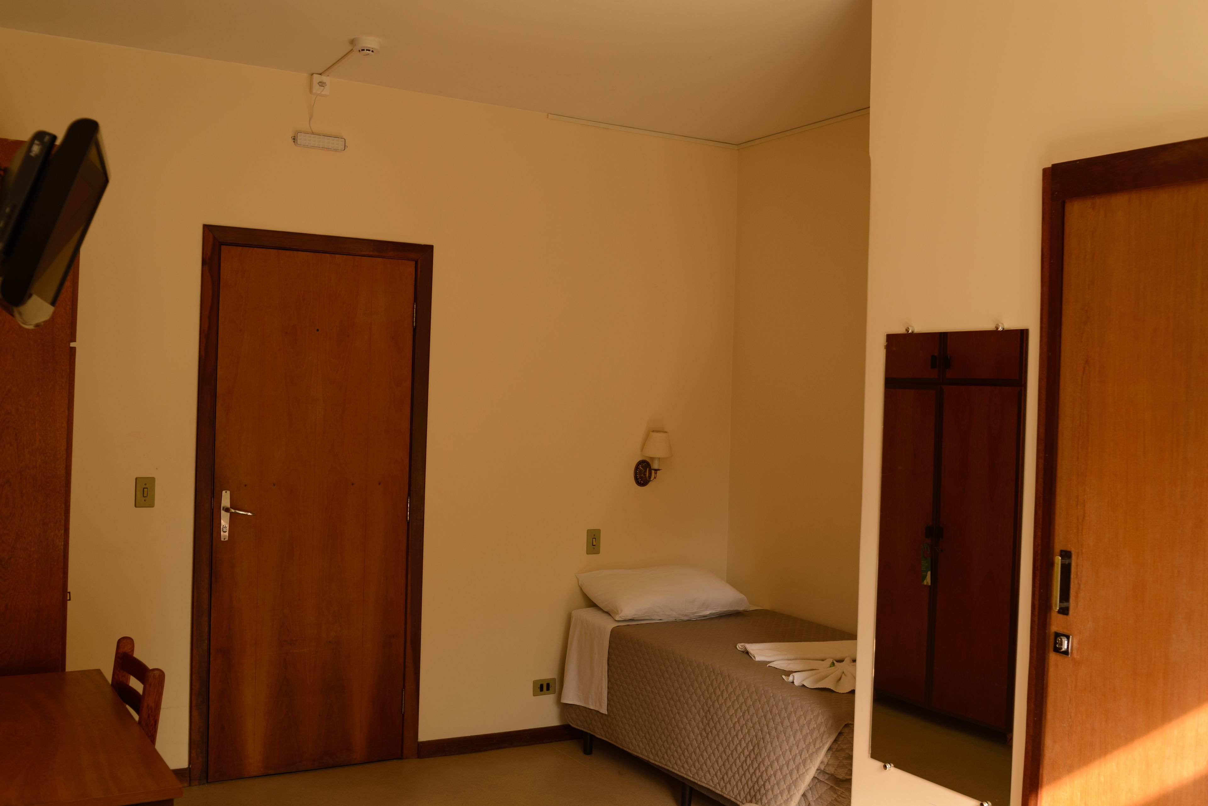 A room with a bed, a wooden door, a wall-mounted light, and a small table.