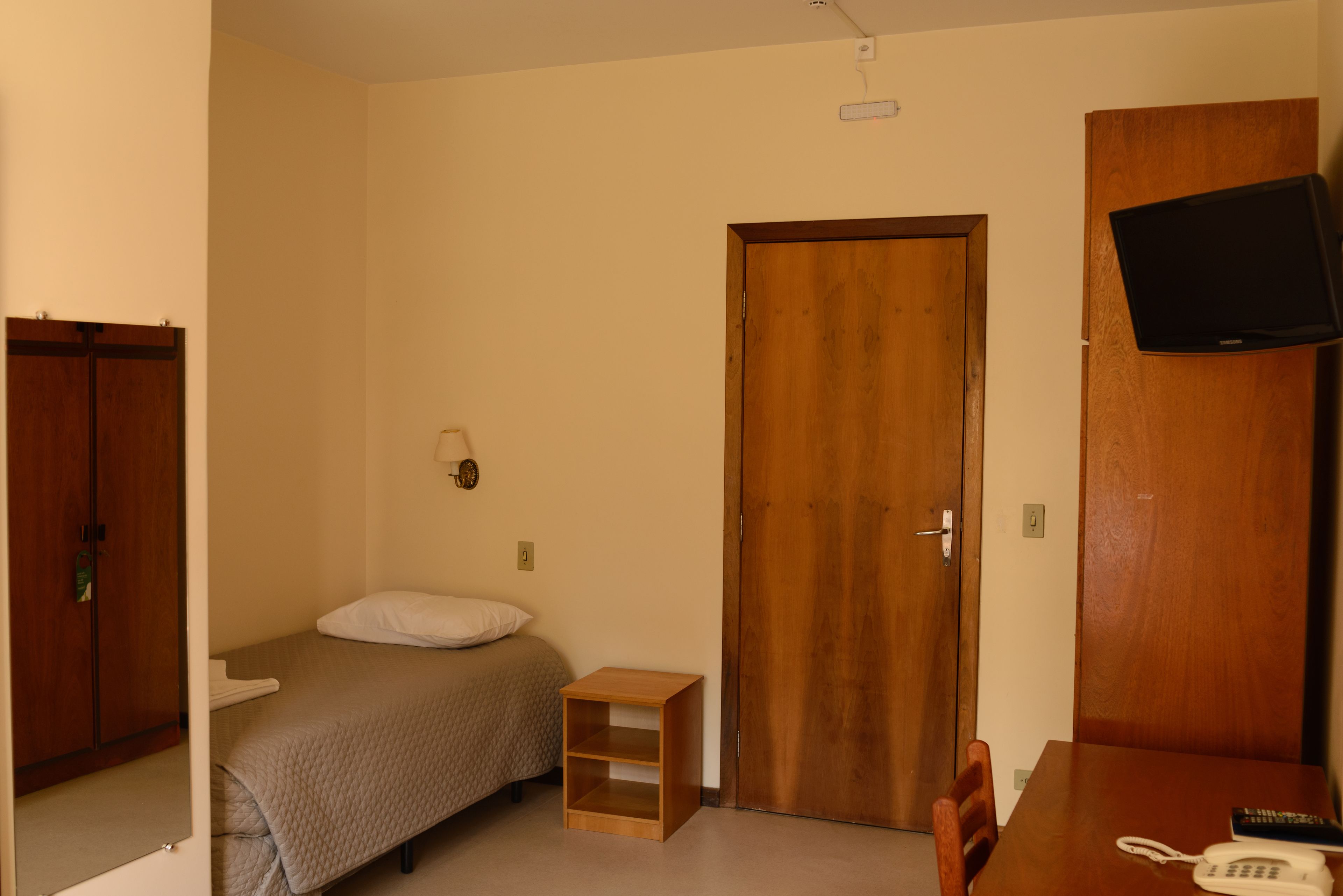 Una habitación de hotel pequeña y de un solo ambiente con cama, una mesita de noche de madera, un escritorio con teléfono y control remoto, un televisor montado en la pared y una puerta.