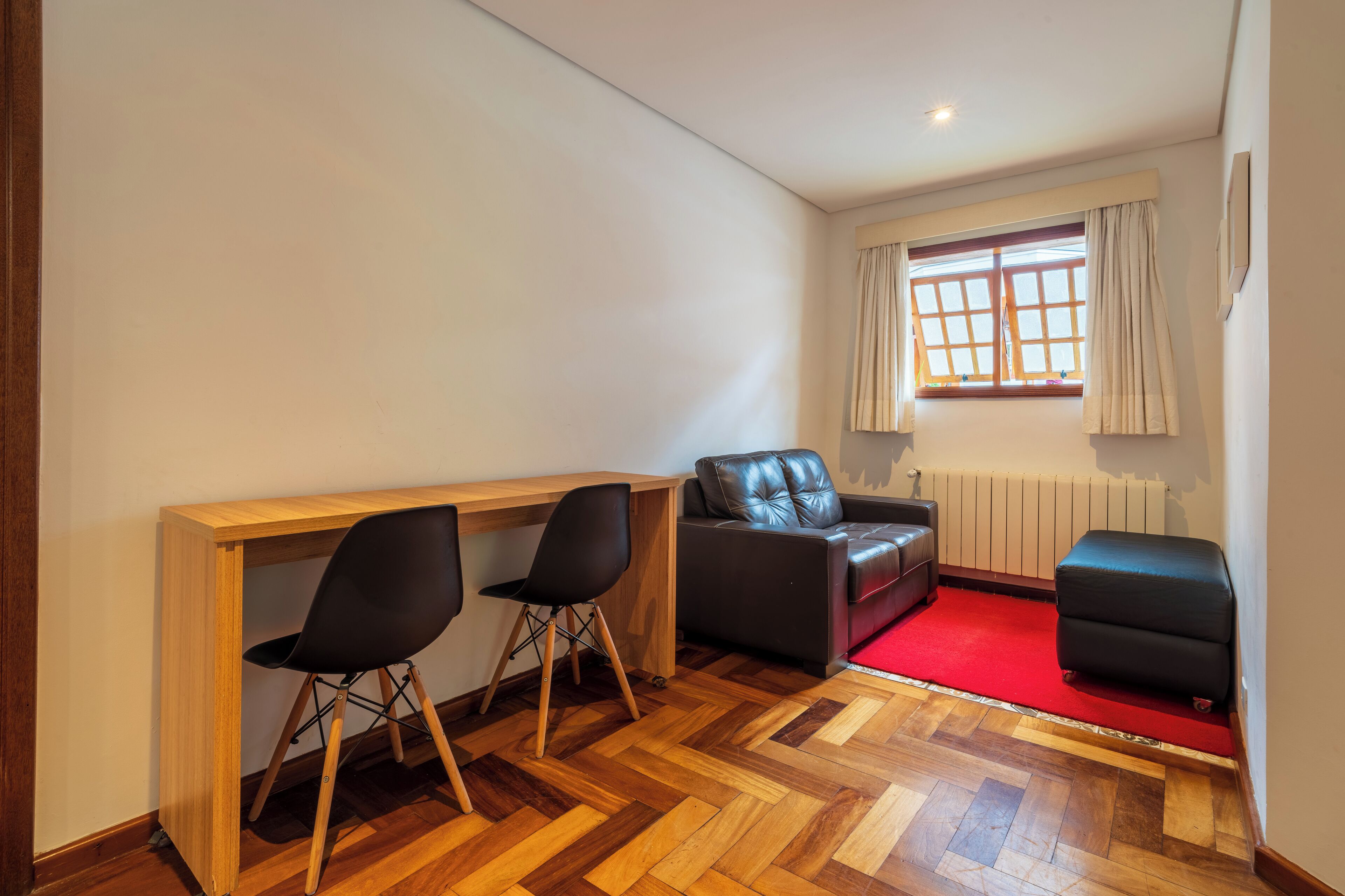 A compact room with a wooden desk, two black chairs, a leather sofa, and a red carpet.